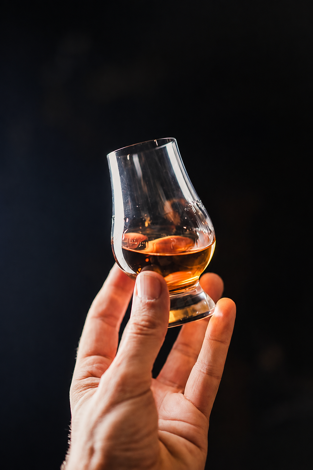 Hand holding a tasting glass of amber American whiskey against a dark background
